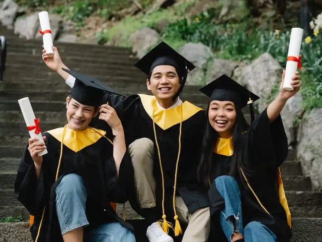 Three graduates sitting on steps, holding up their diplomas